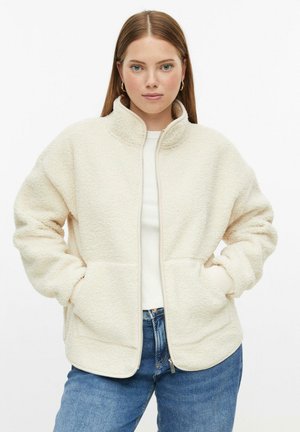 Donna che indossa una giacca in pile color crema con zip, mani in tasca e jeans blu, di fronte a uno sfondo bianco.