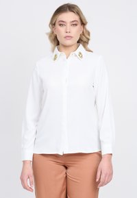 Blusa bianca con chiusura a bottoni sul davanti e colletto decorato con pietre gialle e trasparenti. Maniche lunghe; tessuto liscio con leggero lucido.
