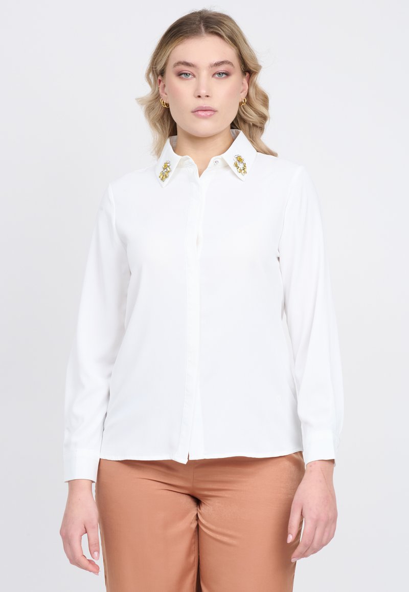 Blusa bianca con chiusura a bottoni sul davanti e colletto decorato con pietre gialle e trasparenti. Maniche lunghe; tessuto liscio con leggero lucido.