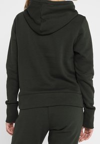 Sudadera de color verde oscuro hecha de un tejido suave. Cuenta con capucha, mangas largas, puños acanalados y dobladillo. Textura lisa con silueta ajustada.