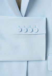 Manche de veste de costume bleu clair avec quatre boutons recouverts du même tissu et revers cranté visible en arrière-plan.