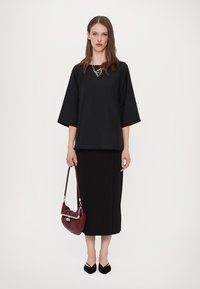 Zwarte top met driekwartmouwen, gecombineerd met een lange zwarte rok. Accessoires met een bordeauxrode tas en zwarte platte schoenen.