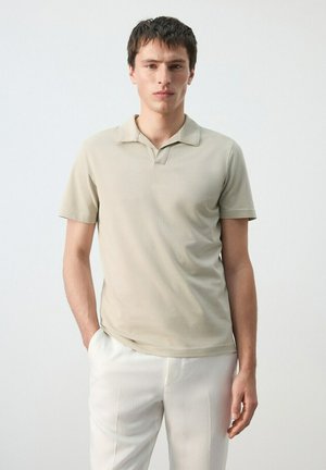 Mladi moški stoji, oblečen v bež polo majico s kratkimi rokavi in bele hlače, z eno roko v žepu na enobarvnem ozadju.