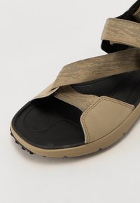 Beige sandals with cross-over fabric straps, textured surface, and a black rubber sole. Features a raised pattern for grip and flexibility.