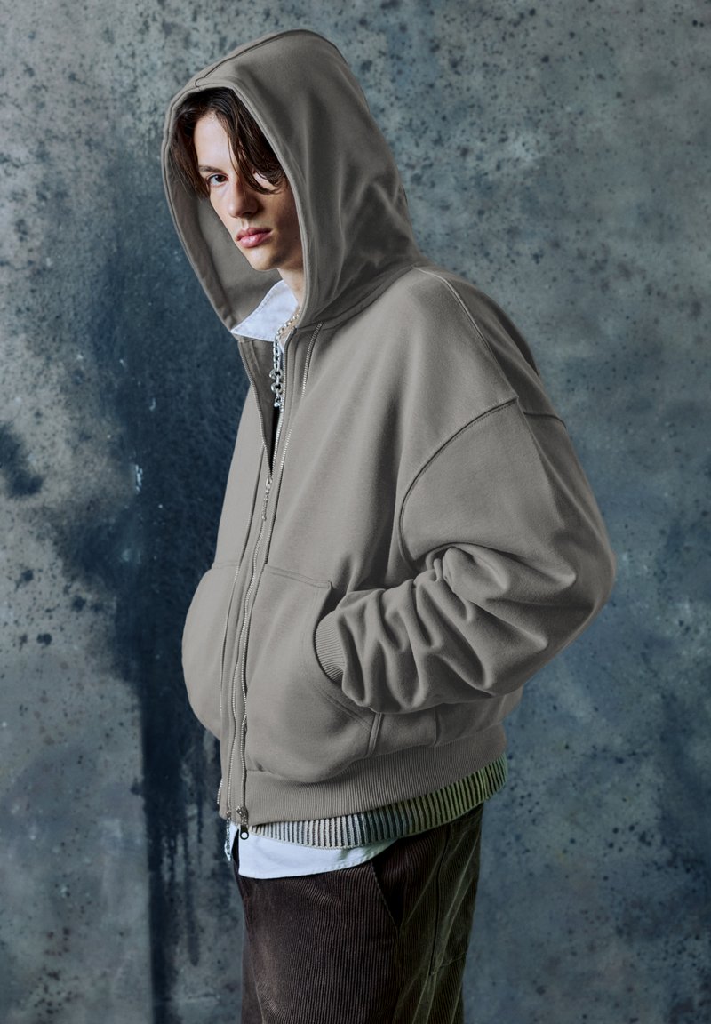 Felpa grigia con zip e cappuccio ampio, dotata di cerniera frontale e tasche laterali. Indossata sopra una camicia bianca e pantaloni di velluto a coste scuri.