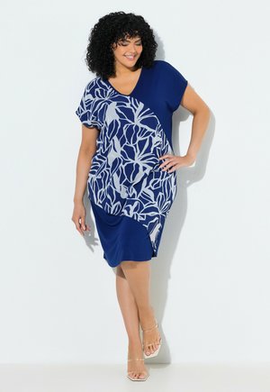Kobieta o kręconych włosach ubrana w niebieską sukienkę do kolan z białymi abstrakcyjnymi wzorami liści, stoi z ręką na biodrze i uśmiecha się.