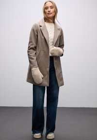 Beżowy płaszcz z mieszanki wełny z szerokimi klapami i dwoma przednimi kieszeniami, noszony na kremowym swetrze w warkocze, zestawiony z ciemnymi dżinsami i rękawiczkami podszytymi futrem.
