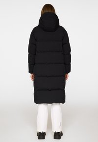 Svart lång puffjacka med huva, med ett kviltat mönster och tydlig vaddering; bärs över vita byxor och svarta stövlar.