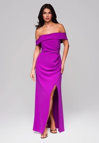 Fialová off-shoulder večerní šaty vyrobené z hladk�ého materiálu, s předním rozparkem a drapérií, doplněné zlatými sandály na kotníku.