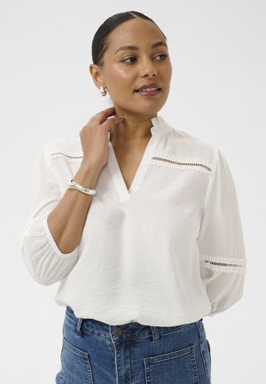 Femme portant un chemisier blanc avec des détails en dentelle et un jean bleu, touchant son cou et regardant sur le côté devant un fond uni.