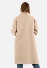 Abrigo de lana beige con un corte relajado, mangas oversized y cuello con muesca. Presenta una textura premium y un diseño de longitud media.