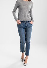 Maglietta grigia chiara a maniche lunghe, jeans in denim blu con vestibilità slim e décolleté metallici a punta. L'outfit presenta tasche e una texture sottile.