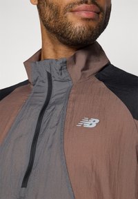 Lätt jacka med hög krage, grå och bruna färgblock, dragkedja fram och New Balance-logotyp på bröstet. Strukturerad tyg.