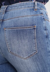 Jeans de mezclilla azul desgastados con cintura media, que cuentan con un bolsillo trasero y detalles de costura visibles. Textura suave con un ajuste ceñido.