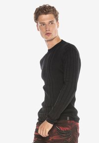 Schwarzer gerippter Strickpullover mit Rundhalsausschnitt. Verfügt über ein strukturiertes Muster und lange Ärmel, kombiniert mit roten gemusterten Hosen.