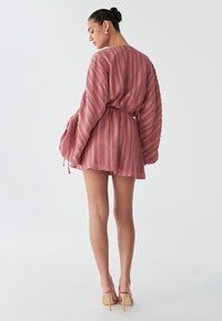 Eine Frau mit einem Dutt trägt ein rosa, gefaltetes Kleid mit einem elastischen Bund und steht vor einem schlichten weißen Hintergrund.