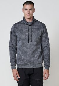 Hombre vistiendo un sudadera con capucha gris jaspeada y un cuello alto, con pantalones cargo negros, de pie contra un fondo claro y liso.