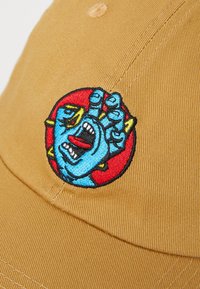 Tan baseballcap med et fargerikt brodert design av et blått ansikt med åpen munn og hevet hånd, satt mot en rød sirkulær bakgrunn.