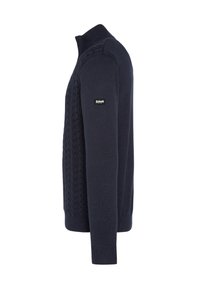 Granatowy, dziergany sweter z wysokim kołnierzem, wzorem w warkocze na przodzie, ściągaczami na mankietach i dole oraz oznaczoną metką na rękawie.