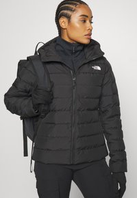 Crna puffer jakna s patentnim zatvaračem i visokim ovratnikom. Ima horizontalne šavove, logotip North Face i uski kroj. Nosi se s rukavicama.