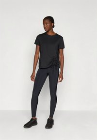 Donna in un top nero arricciato a maniche corte, leggings neri e sneakers nere in piedi davanti a uno sfondo bianco semplice.