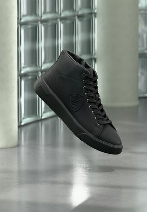 Sneaker alte in pelle nera con lacci e logo inciso, che fluttuano sopra un pavimento in cemento lucido vicino a pareti in blocchi di vetro.
