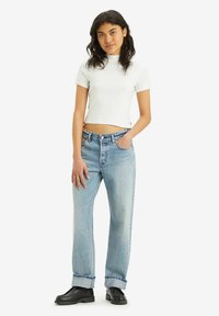 Witte cropped t-shirt, lichtblauwe rechte jeans met een hoge taille, opgerolde mouwen en zwarte chunky schoenen, staand tegen een effen achtergrond.