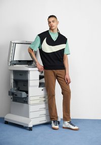 Schwarzer ärmelloser Strickpullover mit weißem Nike-Logo über einem mintgrünen Shirt, kombiniert mit braunen Hosen und weißen Turnschuhen auf einem blauen Boden.