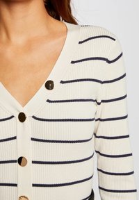 Donna che indossa un cardigan rigato crema con strisce orizzontali nere e bottoni rotondi dorati, che mostra la parte superiore del torso e il collo.