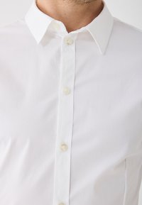 Férfi fehér ingben, hegyes gallérral és gombos elülső részen, látható nyakkal és felső mellel.