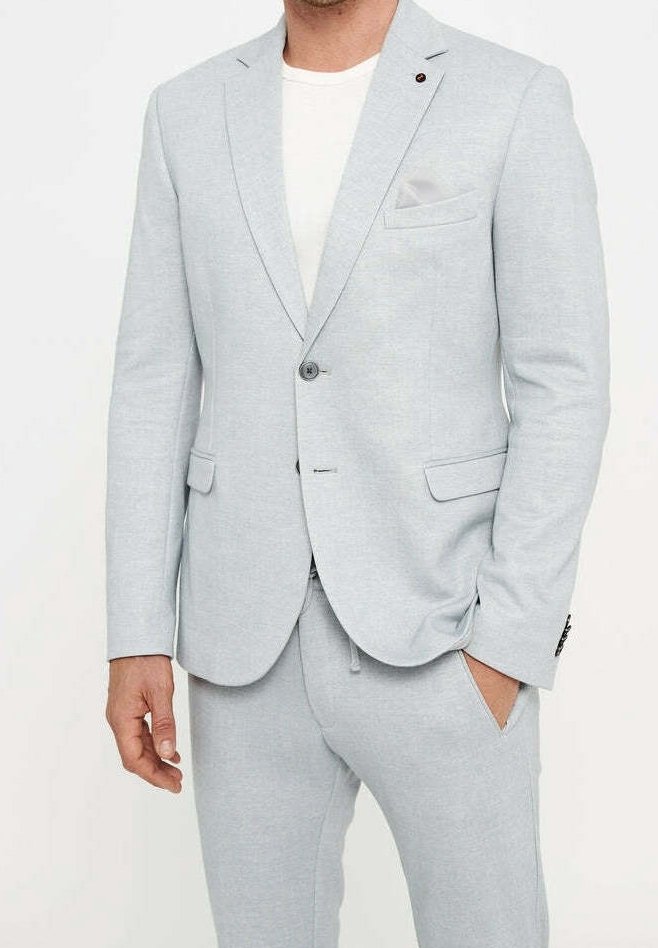Homme portant une veste de costume ajustée gris clair et un pantalon assorti avec une chemise blanche en dessous, une main dans la poche.