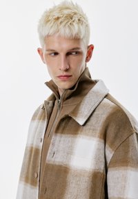 Ung man med kort platinablont hår, iklädd en beige och vit rutmönstrad jacka över en brun muddtröja med dragkedja, tittar mot sin vänstra sida.