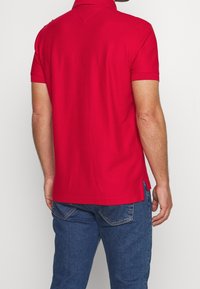 Červené polo tričko s krátkými rukávy, límečkem a postranními rozparky; vyrobené z hladké tkaniny. Zadní strana má jednoduchý design bez vzorů.