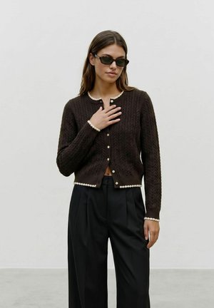 Femme portant un cardigan noir texturé avec une bordure blanche, un pantalon noir taille haute et des lunettes de soleil foncées, se tenant devant un fond clair uni.