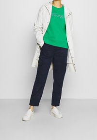 Sweatshirt vert avec logo brodé, superposé sous une veste de pluie blanche, associé à un pantalon cargo bleu marine et des baskets blanches.
