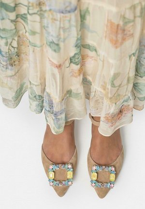 Pies con bailarinas puntiagudas color canela con hebillas de joyas coloridas, parcialmente cubiertos por una falda transparente de flores con estampado floral.