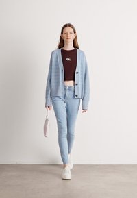 Svetlomodrý rebrovaný kardigán cez crop top s logom, doplnený svetlomodrými obtiahnutými džínsami a bielymi teniskami. Drží malú ružovú tašku.