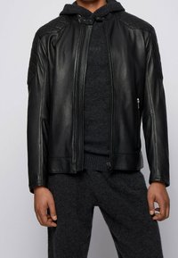 Černá kožená bunda s prošívanými akcenty na ramenou, zapínáním na zip a mikinou pod ní. Texturovaná tkanina, přiléhavý střih a přední kapsy.