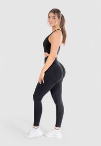 Czarny, bezszwowy sportowy crop top i legginsy, cechujące się dopasowanym krojem, gładką teksturą oraz kontrastowym białym logo marki na pasku w talii.