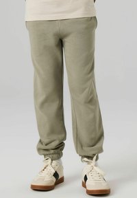 Jasnozielone spodnie dresowe ze ściągaczami, o gładkiej fakturze. Połączone z białymi sneakersami z czarnym paskiem i brązowymi podeszwami.