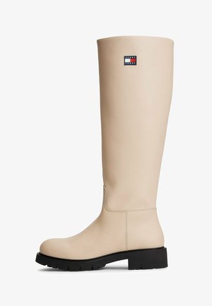 Beige knæhøje læderstøvle med sort sål og lille Tommy Jeans-logo nær toppen på ydersiden.