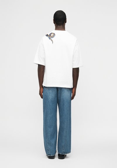 Άνδρας με λευκό φαρδύ T-shirt με γραφικό φίδι στον ώμο, άνετο μπλε τζιν και μαύρα παπούτσια, στέκεται μπροστά σε απλό λευκό φόντο.