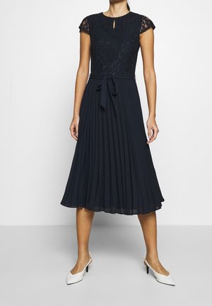 Kvinde iført sort blonderkjole med plisseret nederdel og bundet talje, kombineret med hvide lavhælede slingback-sko, står foran en ensfarvet baggrund.