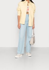 Personne portant une chemise boutonnée jaune pâle, un t-shirt graphique, un jean à jambe large bleu clair, des baskets beiges, et tenant un sac à main beige avec une bandoulière chaîne.