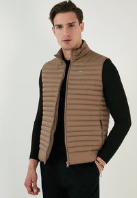 Vesta puff din material maro, cu cusături verticale, guler înalt și buzunare cu fermoar. Poartă peste o cămașă neagră cu mâneci lungi. Textură netedă.