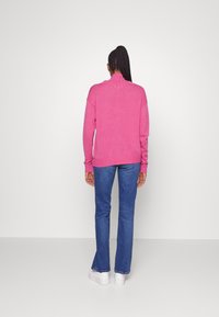 Roze gebreide trui met een neklap en ribgebreide manchetten, gecombineerd met flare blauwe jeans en witte sneakers, bekeken vanaf de achterkant.