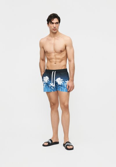 Homme torse nu portant un short de bain bleu à imprimé palmiers et des sandales noires Moschino, debout les mains dans les poches devant un fond uni.