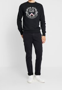 Sort sweatshirt med logo og våbenskjold-design, kombineret med mørke bukser og hvide sneakers. Afslappet pasform, ribstrikkede manchetter og kant.