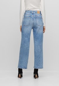 Nainen, joka on pukeutunut vaaleansinisiin suoriin farkkuihin ja mustiin korkokenkiin, seisoo yksinkertaista vaaleaa taustaa vasten, selkä katsojaan päin.