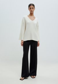 White ribbed sweater with a V-neck and wide sleeves, paired with black flared trousers and flat sandals, set against a plain background.